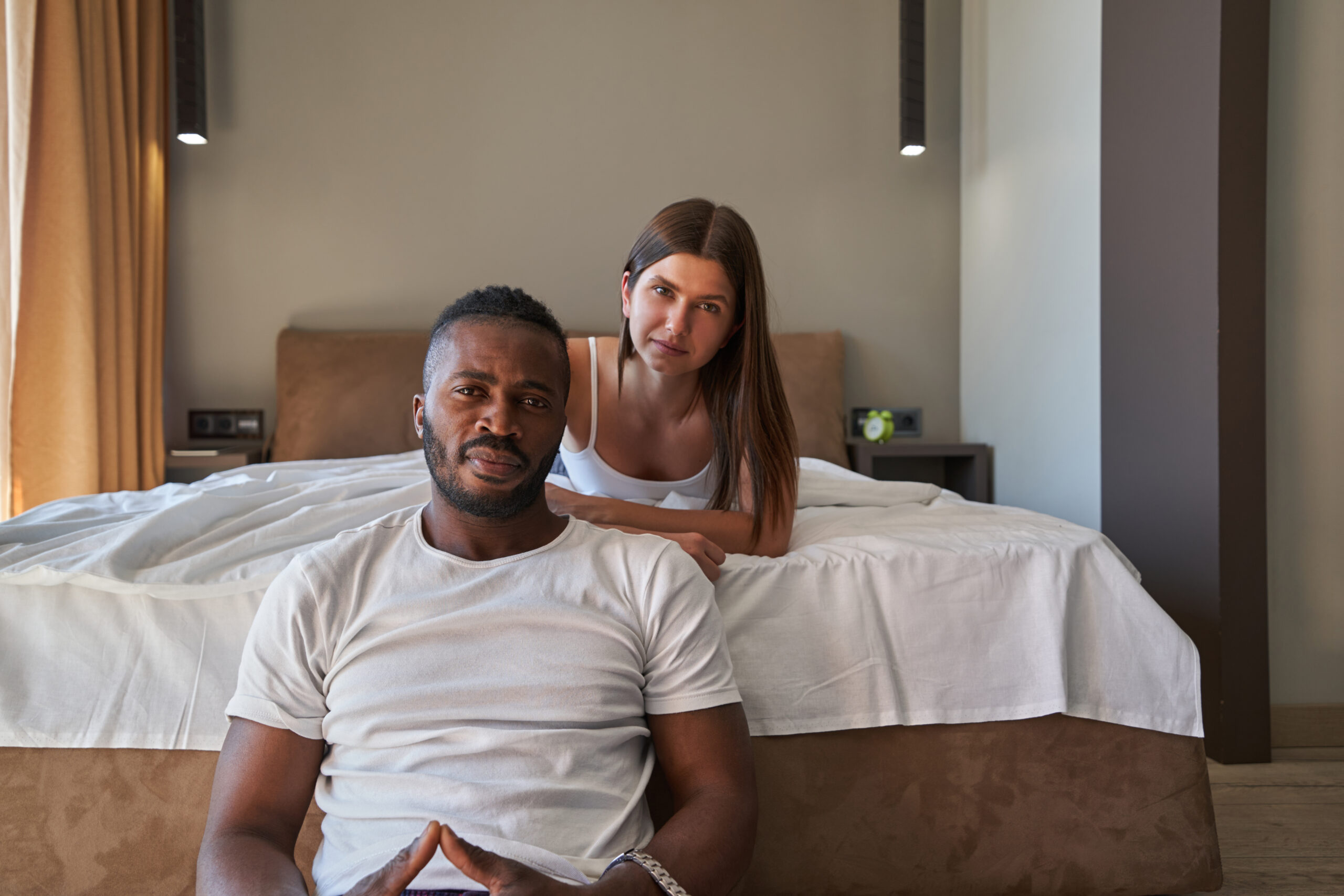 Interracial couple sitting together in a bedroom, looking thoughtful and serious, representing the emotional challenges and relationship strain couples faced during COVID-19 lockdowns.